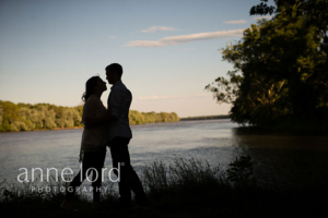 An couple celebrate their engagement on a lake with a setting sun. Photograph by Anne Lord Photography